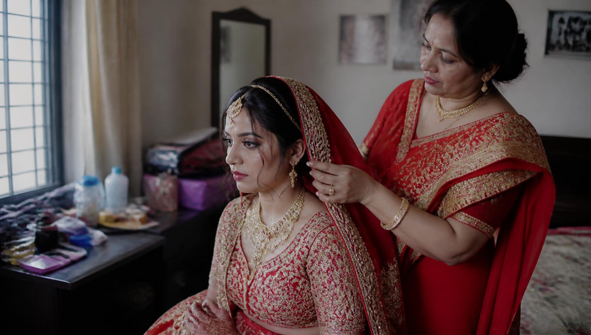 Bride sitting by a window while her mother adjusts her dupatta